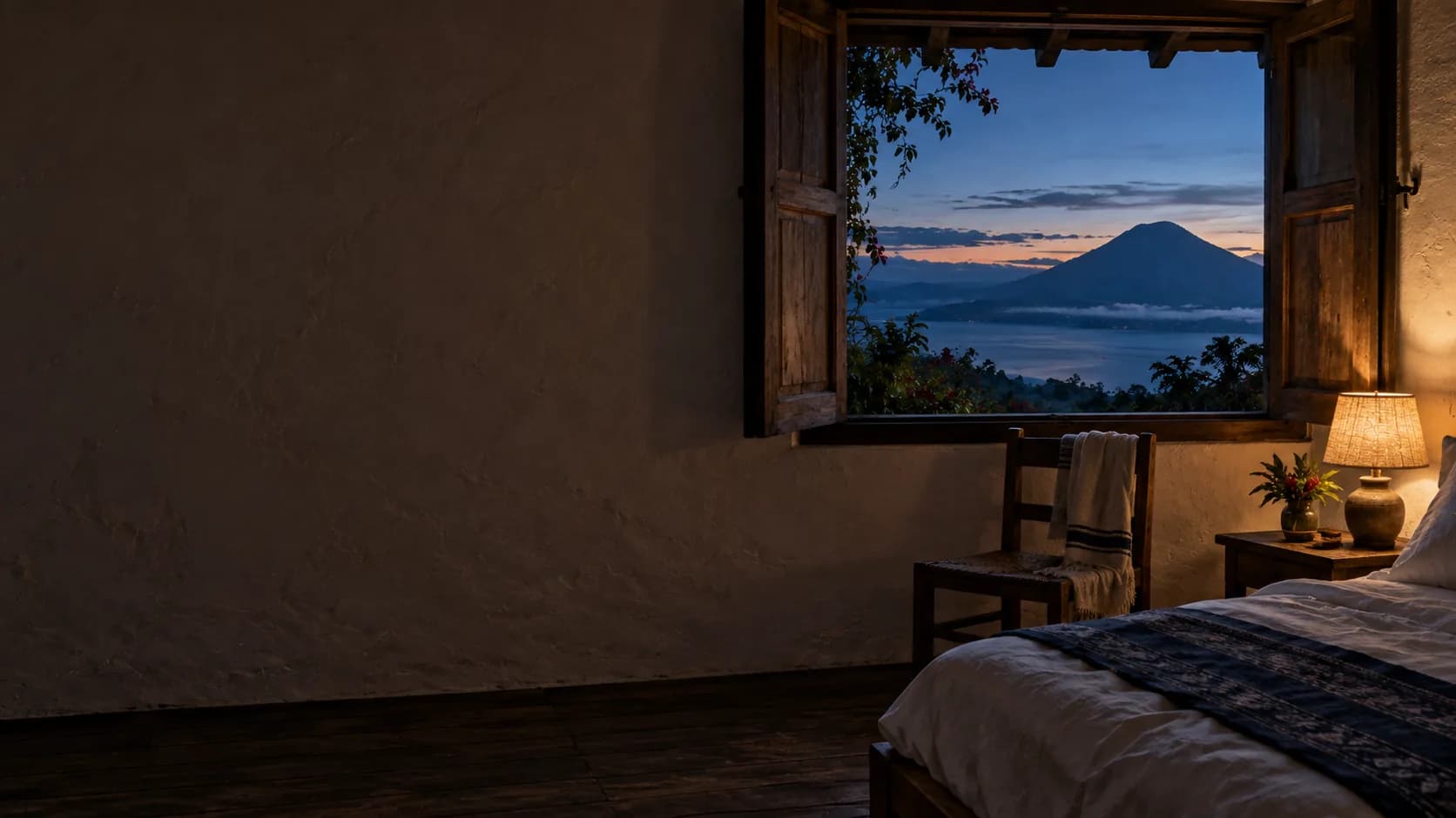 A guest room at dusk, the window open to the lake and one lamp lit.